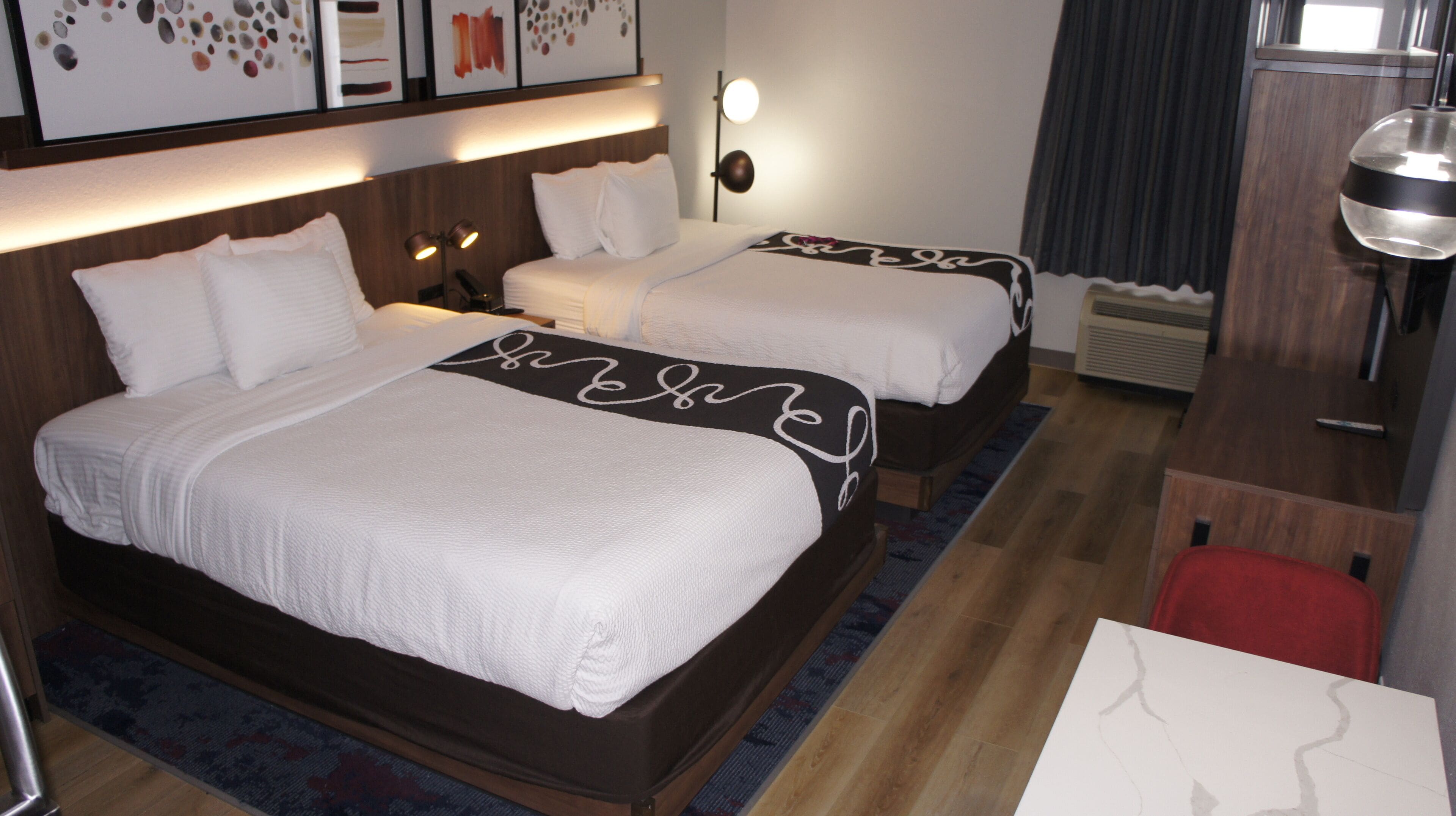 A hotel room with two beds, a desk, and a chair. There are wall-mounted lamps and a heater.