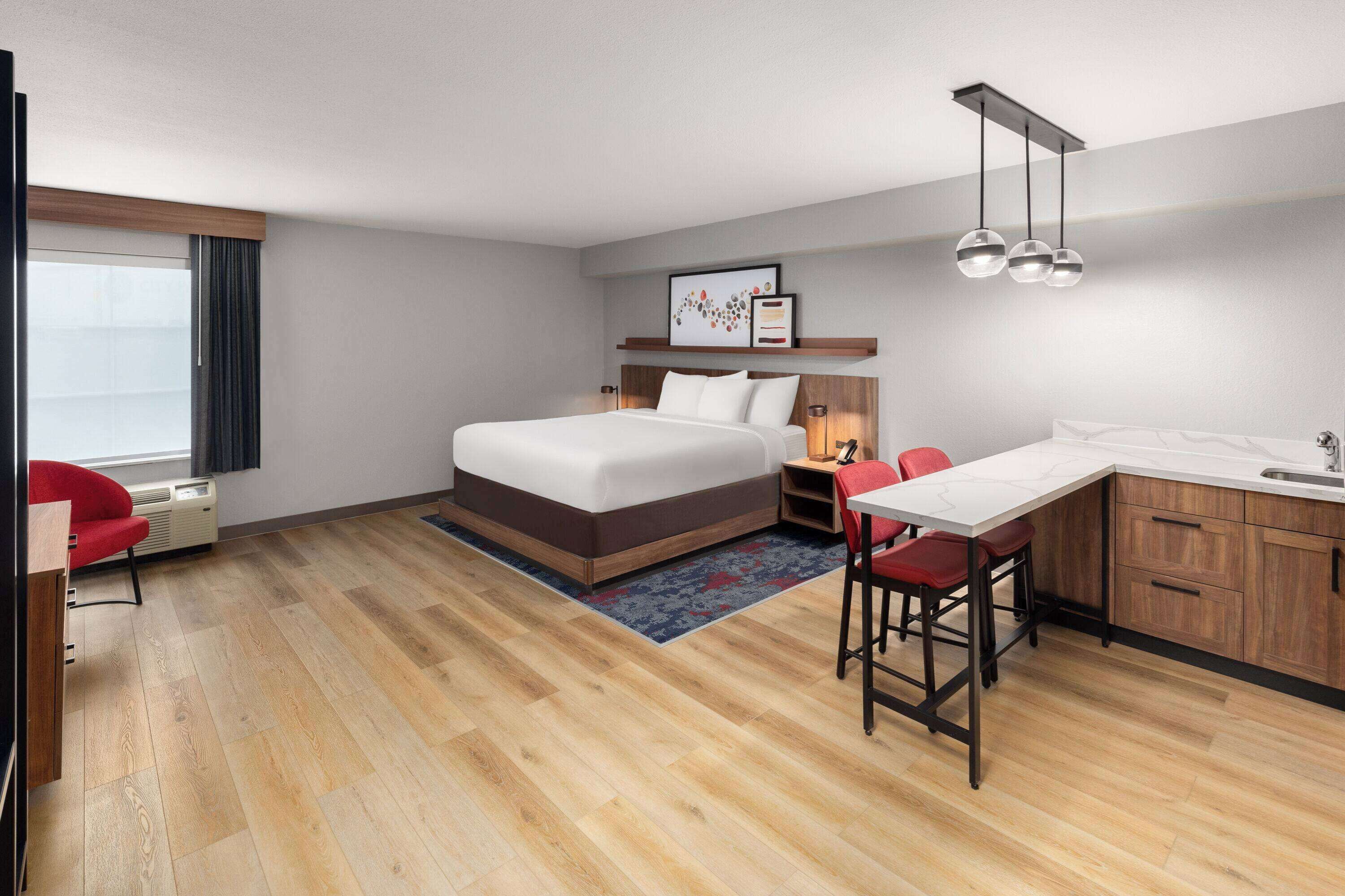 A modern hotel room with a bed, a desk, a chair, a small kitchen area, and a window with curtains.