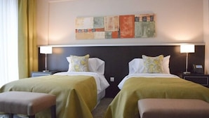 Quarto de hotel com duas camas, cada uma com travesseiro e mesa de cabeceira. Há um quadro na parede acima das camas.