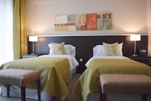 A hotel room with two beds, each with a pillow and a bedside table. There is a painting on the wall above the beds.