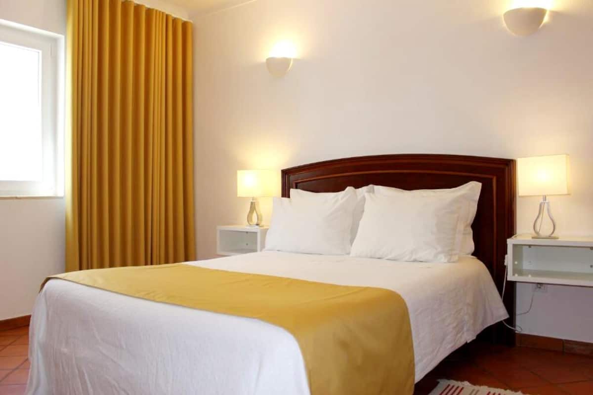 A neatly made bed with white bedding and a yellow throw, two bedside lamps, and a window with curtains.