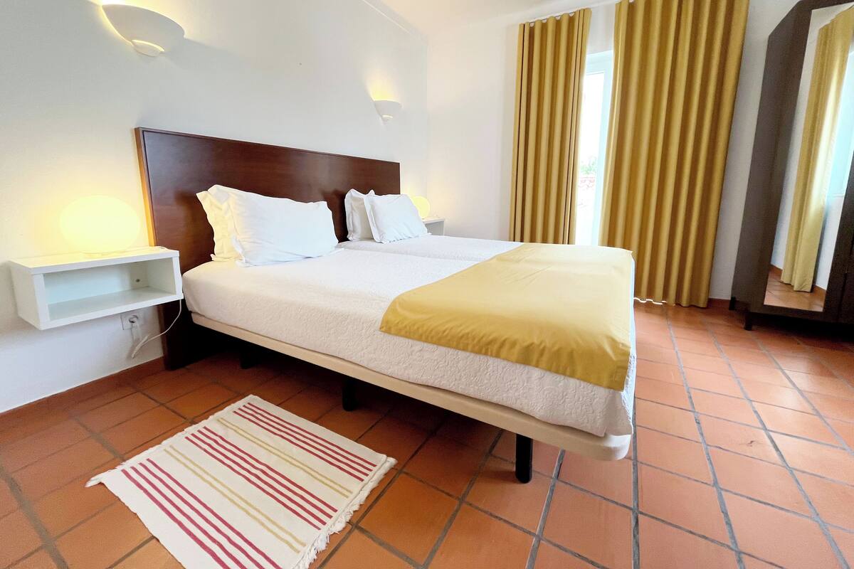 A hotel room with a bed, a bedside table, a lamp, curtains, and a tiled floor.