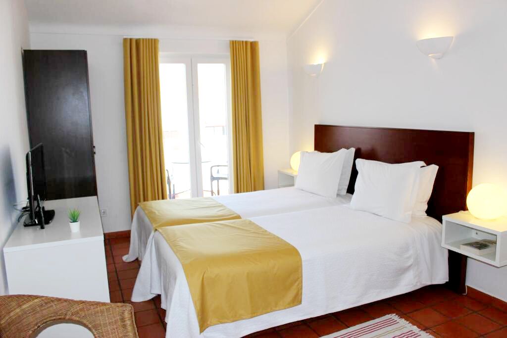 Ein Hotelzimmer mit einem Doppelbett, zwei Nachttischen, einem Fernseher und einem kleinen Schreibtisch mit einer Pflanze.