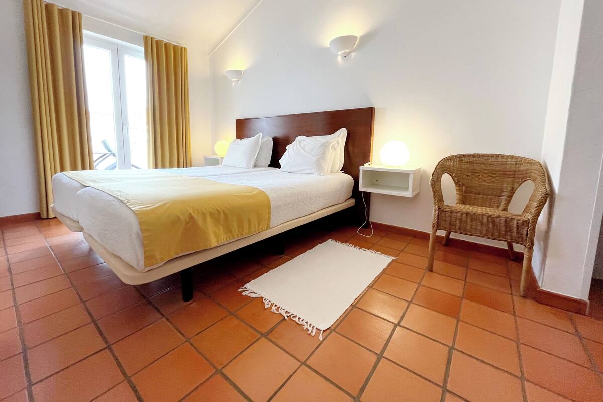 A hotel room with a bed, a nightstand, a chair, and a wall-mounted lamp.