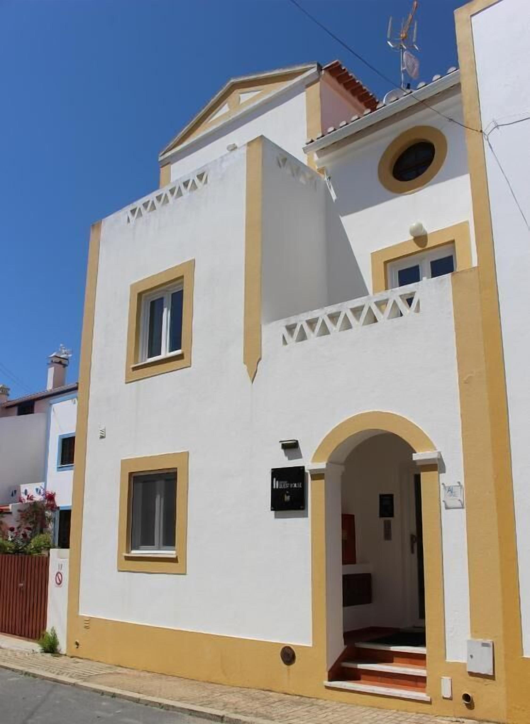 Ein zweigeschossiges Gebäude mit weißer Fassade und gelbem Zierdekor. Ein Schild weist auf ein Geschäft oder eine Dienstleistung im Erdgeschoss hin.