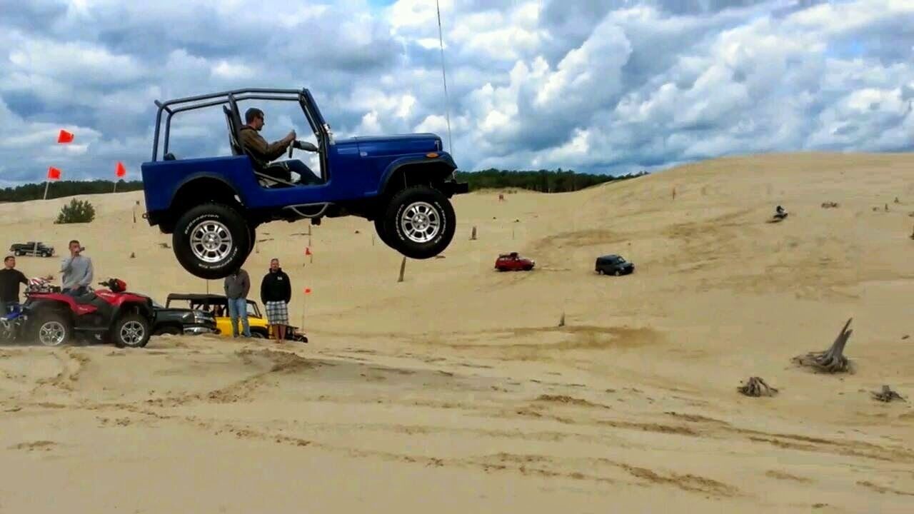 Un vehículo todoterreno azul en el aire, rodeado de gente y otros vehículos sobre terreno arenoso.