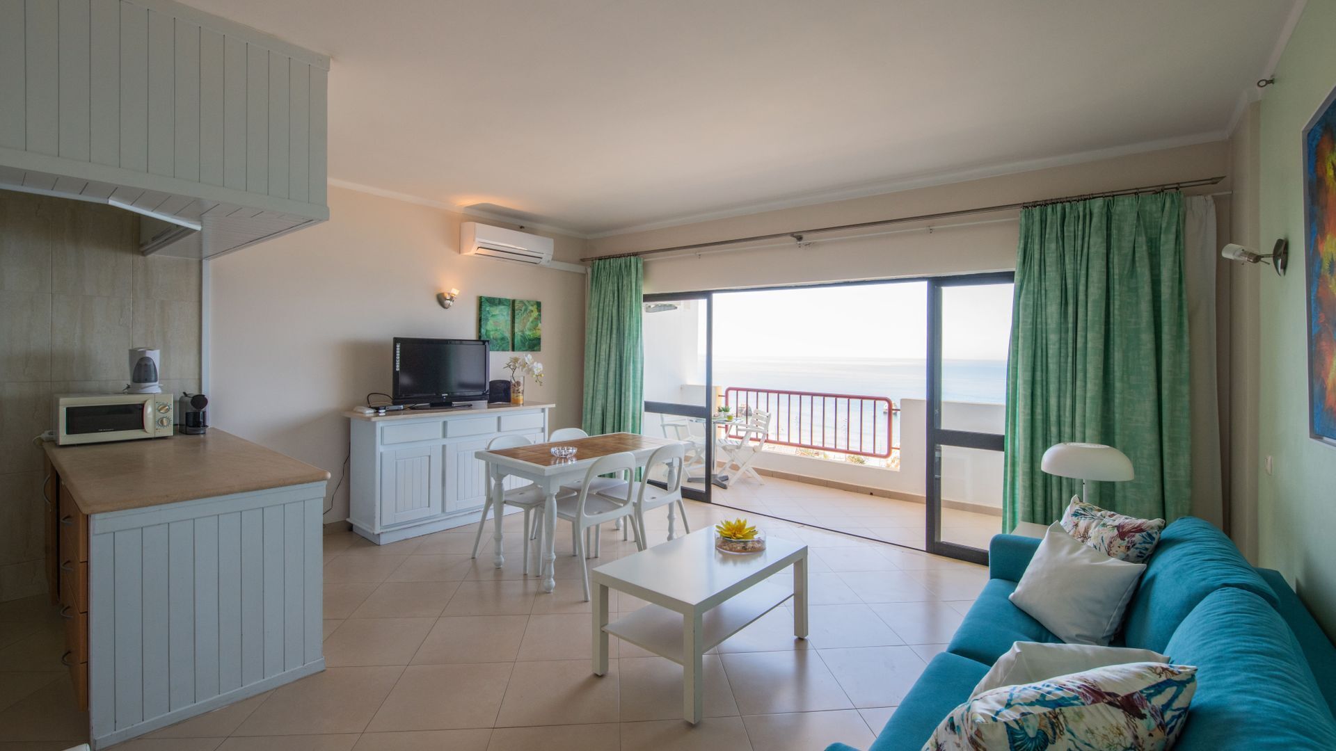 A modern living room with a kitchen, a dining area, and a balcony with a view of the sea.