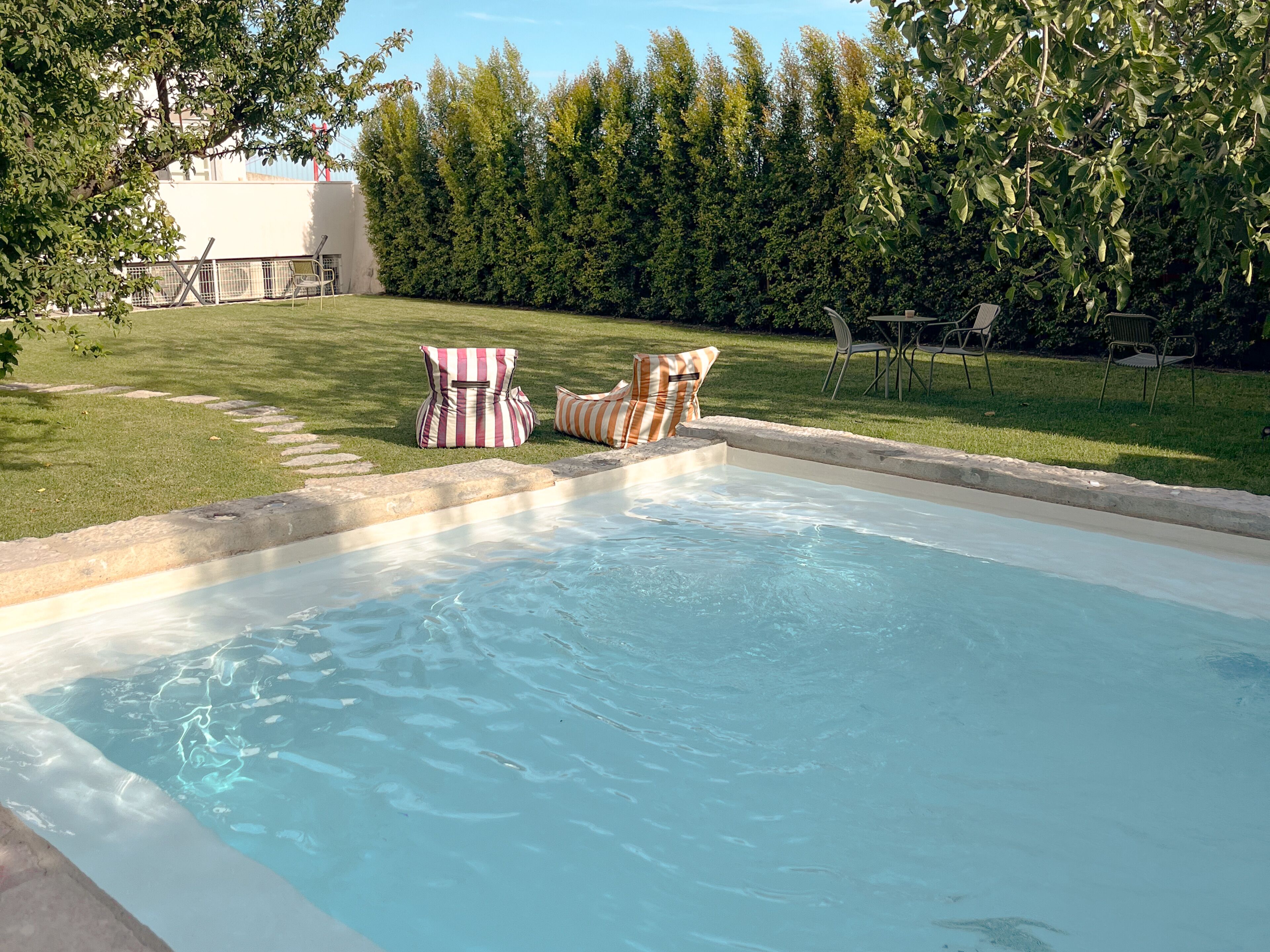 Piscina com duas espreguiçadeiras listradas e área gramada com cerca e árvores.