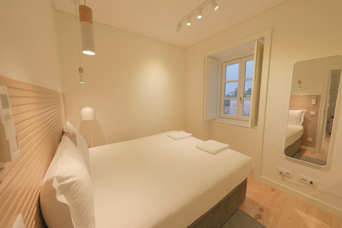A neatly made bed with white linens, a bedside lamp, and a window with a view of the outside.