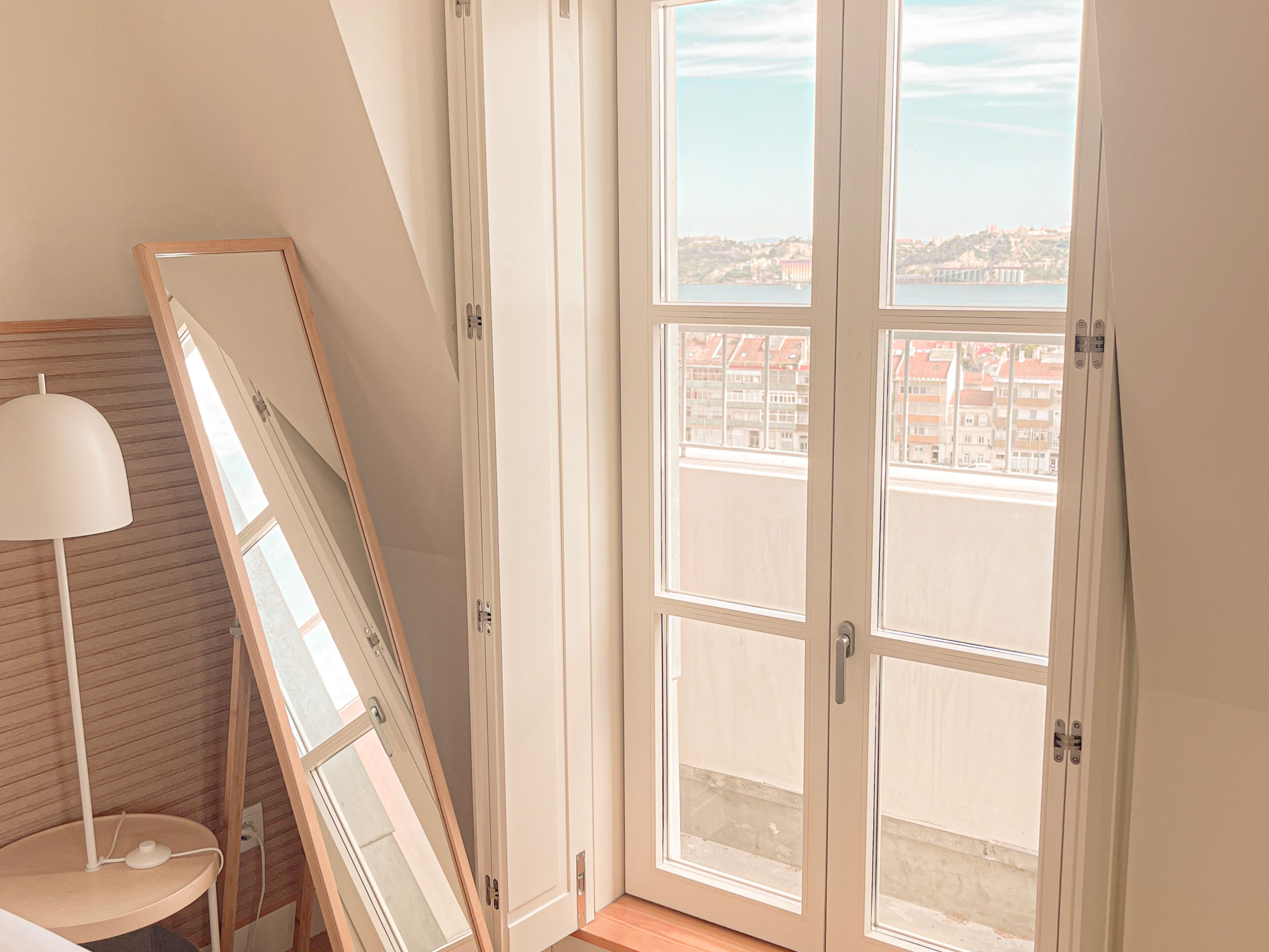 A room with a large window, a mirror leaning against the wall, and a lamp on a small table.