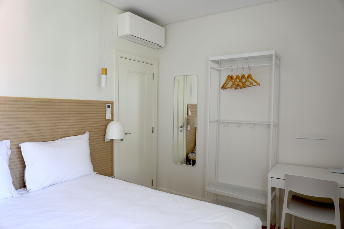 A hotel room with a bed, a desk, a chair, a mirror, and a wardrobe with hangers.