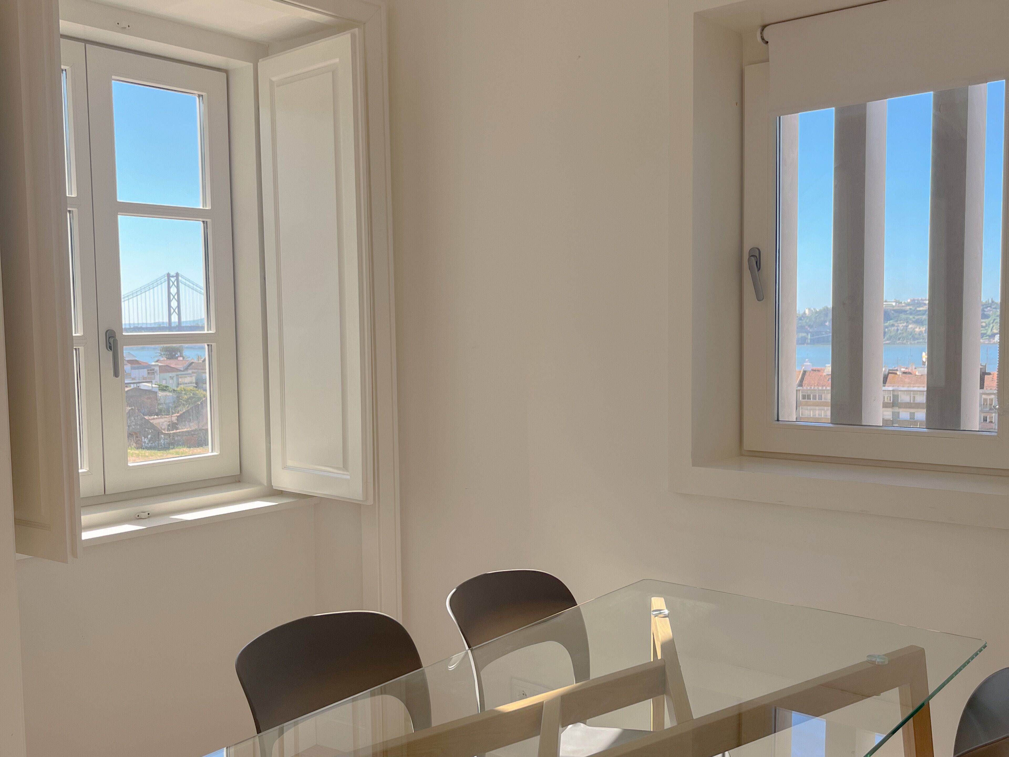 Habitació amb taula i cadires de vidre, finestra amb vista a un pont i edificis, i una porta amb pom.