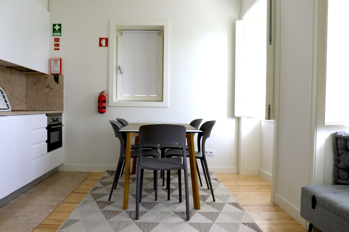Una cocina moderna con armarios blancos, una mesa de comedor con cuatro sillas, un extintor y una ventana.