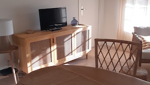 Un salon avec un téléviseur posé sur un meuble en bois, une table à manger avec un set de table en fibres naturelles et un panier de fruits sur la table.