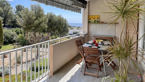 Un balcon avec une table et des chaises, une jardinière suspendue, et une vue sur la mer et les arbres.
