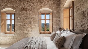 Een slaapkamer met een stenen muur, drie gewelfde ramen, een bed met kussens en uitzicht op het landschap.