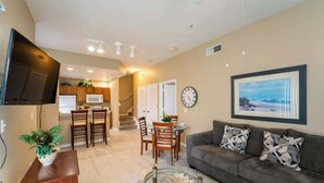 A living room with a sofa, a coffee table, a TV, a dining area with chairs and a table, and a kitchen in the background. - Disney Area Condo w/ 4 Pools & Arcade Fun (Kissimmee)