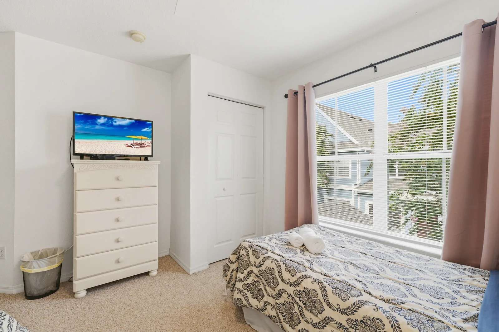 A bedroom with a bed, a dresser, a television on top of the dresser, and a window with curtains.