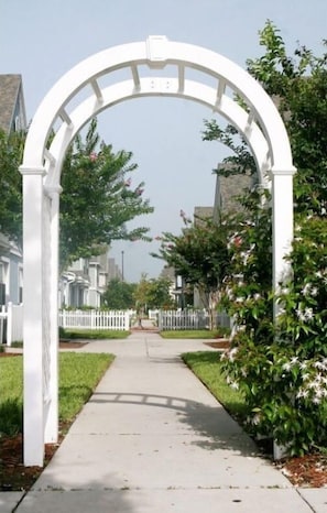 A white arched pathway leading to a gated entrance, surrounded by well-maintained lawns and residential buildings.