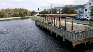 A wooden pier extending over calm water, with a covered seating area and a view of residential buildings and trees in the background.