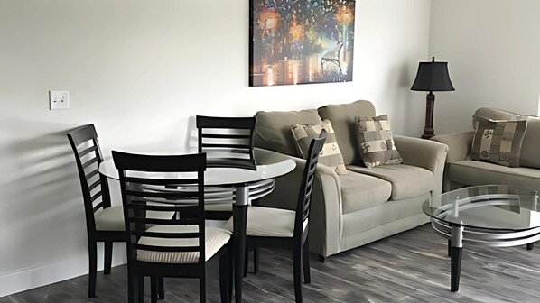Una sala moderna con una mesa redonda de comedor, dos sillas negras, un sofá beige con cojines estampados y una mesa de centro de vidrio.