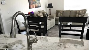 Une cuisine équipée d’un évier, d’une table à manger et de chaises. On aperçoit un canapé et une lampe au fond de la pièce.
