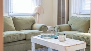 Un salón con dos sofás verdes, una mesa de centro blanca, un libro titulado 'Surf Shack' y una lámpara de pie.