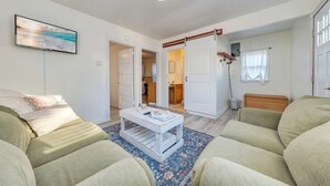 Una sala de estar con sofá, una mesita de centro y un televisor montado en la pared. Hay una puerta que da a otra habitación.