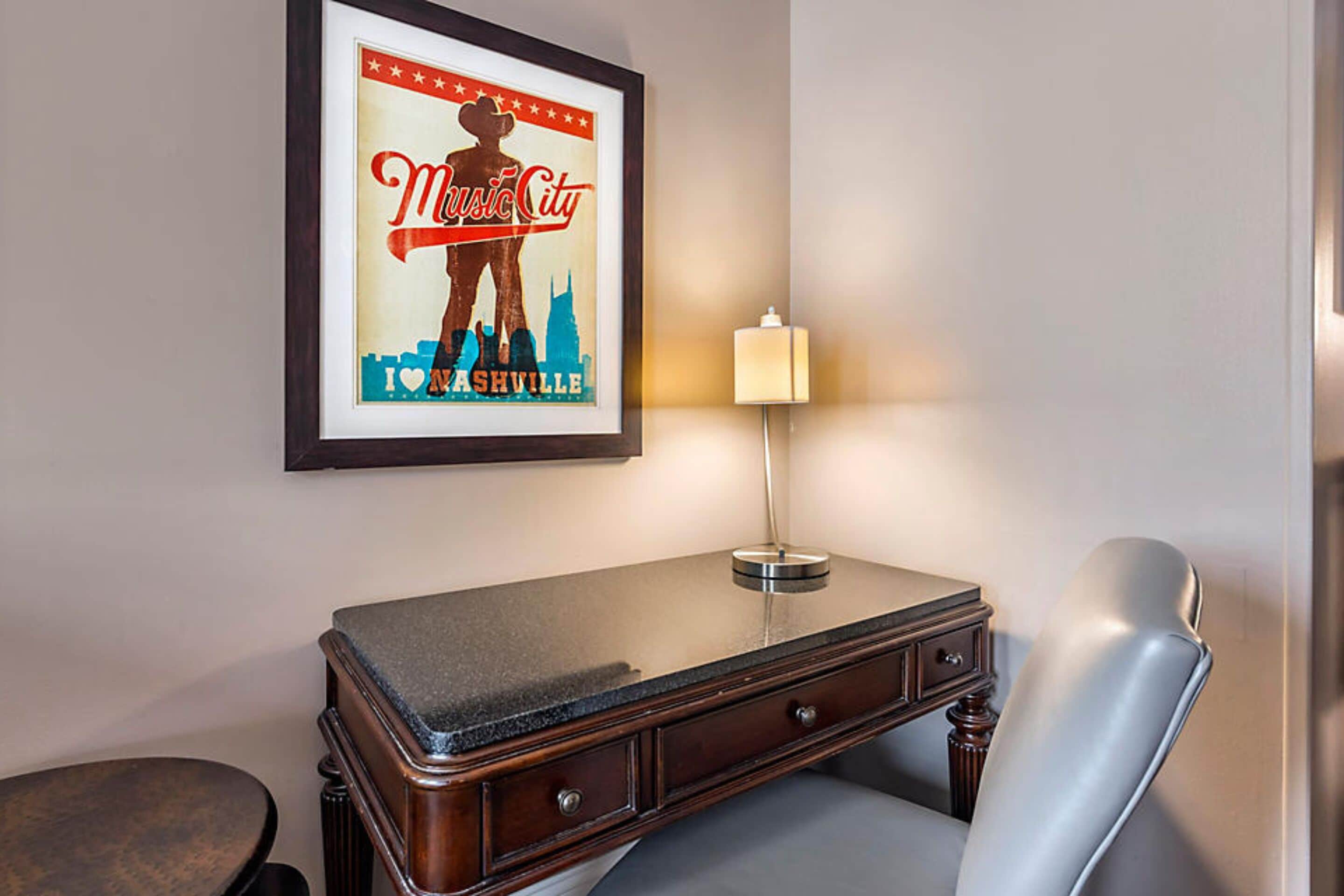 A framed poster of a cityscape with a silhouette of a person and the text "I love Nashville." A desk with a lamp and a chair is in front of the poster.