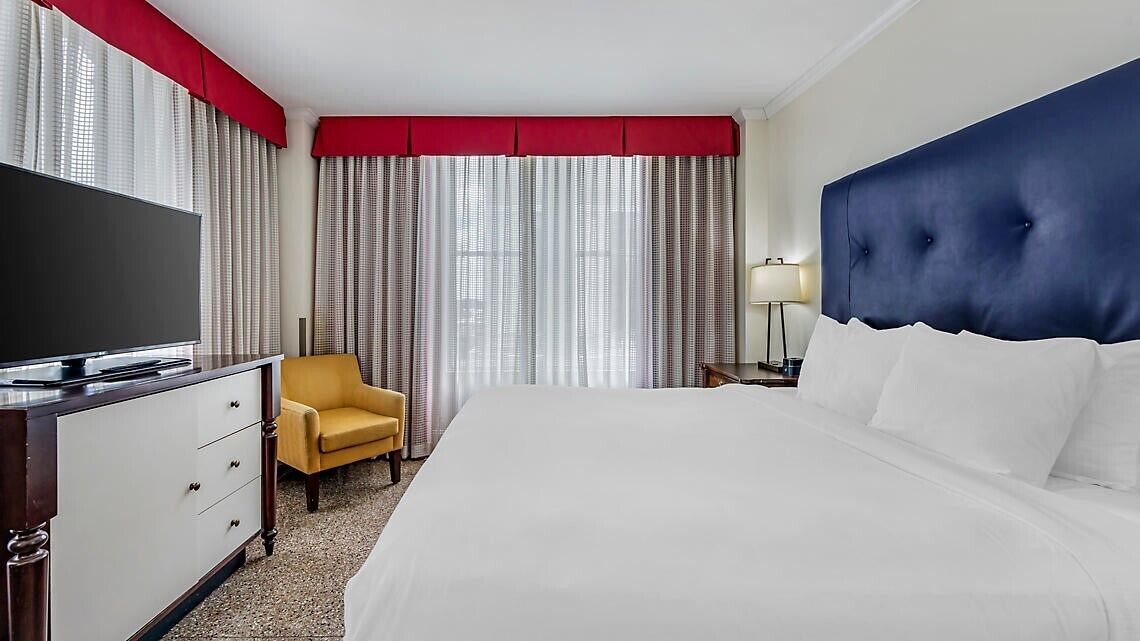 Une chambre d’hôtel avec un grand lit, une télévision, un fauteuil jaune, un bureau avec une lampe et une fenêtre avec des rideaux.