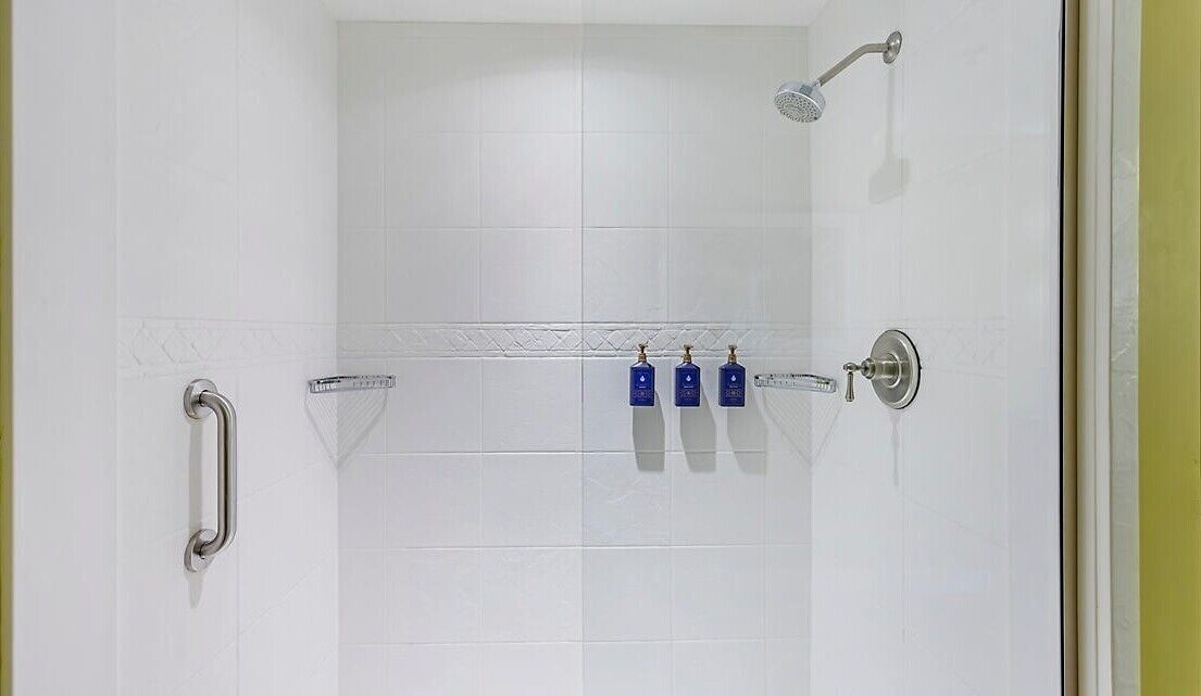 Une salle de bain carrelée de carreaux blancs, une douche avec un pommeau de douche amovible, une barre d’appui et trois bouteilles bleues posées sur une étagère.