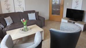 Un salon avec un canapé, une table basse et des fauteuils. Un téléviseur est posé sur un meuble et il y a une porte avec un panneau en verre dépoli.