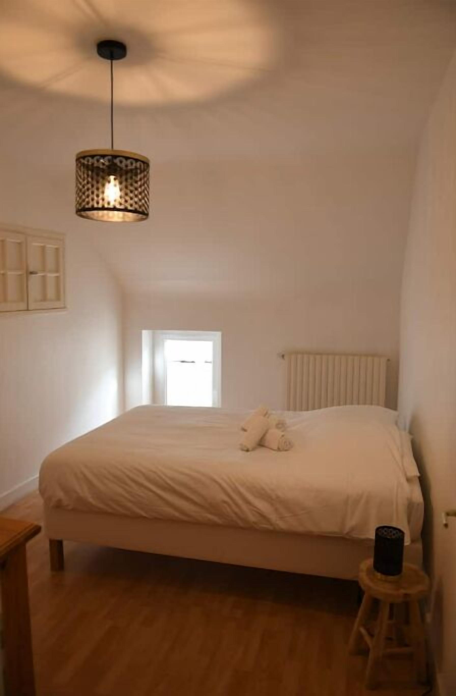 Een slaapkamer met een bed, een klein krukje, een nachtkastje, een raam en een plafondlamp.