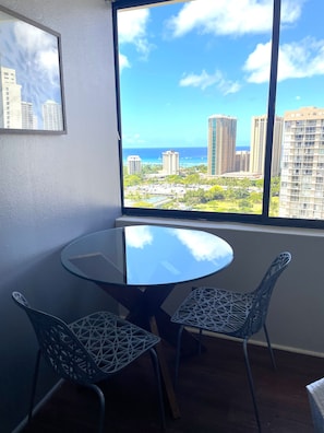 A room with a round glass table and two chairs, a window with a view of buildings and the ocean.