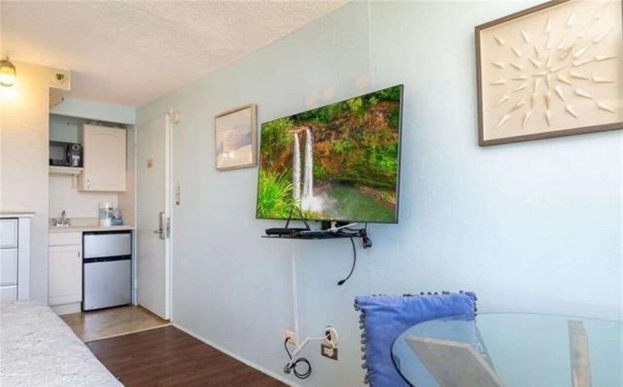 Una sala de estar moderna con televisor montado, una pequeña cocina y una mesa de vidrio con cojín azul.