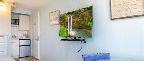 Una sala de estar moderna con televisor montado, una pequeña cocina y una mesa de vidrio con cojín azul.
