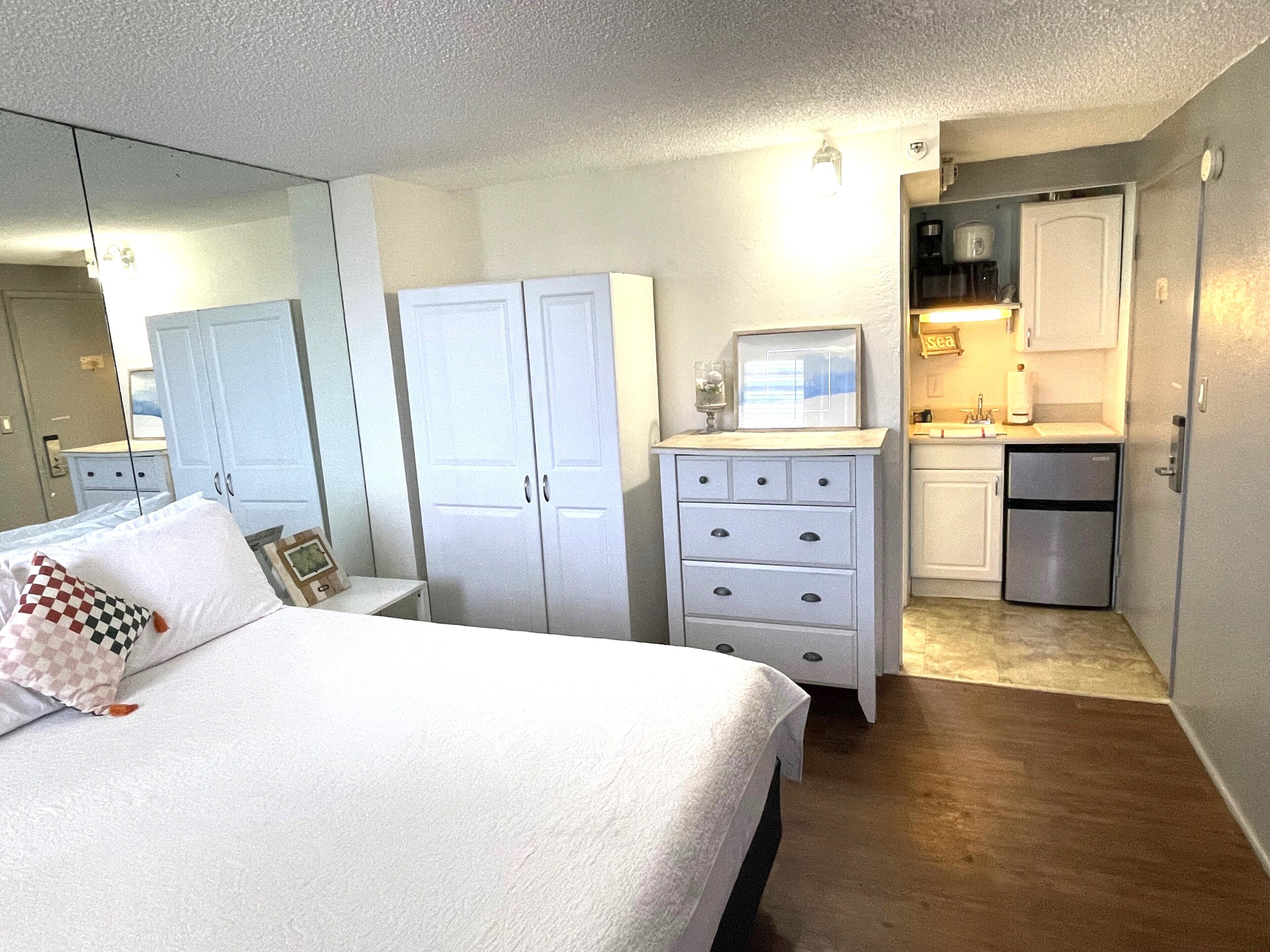A bedroom with a bed, a dresser, a mirror, and a kitchenette with a refrigerator.