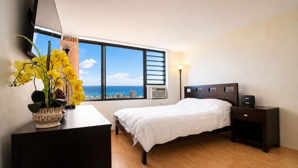 A bedroom with a bed, bedside tables, a desk with a lamp, a potted plant, and a window with a city view. - New Listing Ocean View Studio (Honolulu)