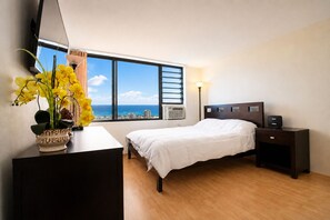 A bedroom with a bed, bedside tables, a desk with a lamp, a potted plant, and a window with a city view. - New Listing Ocean View Studio (Honolulu)