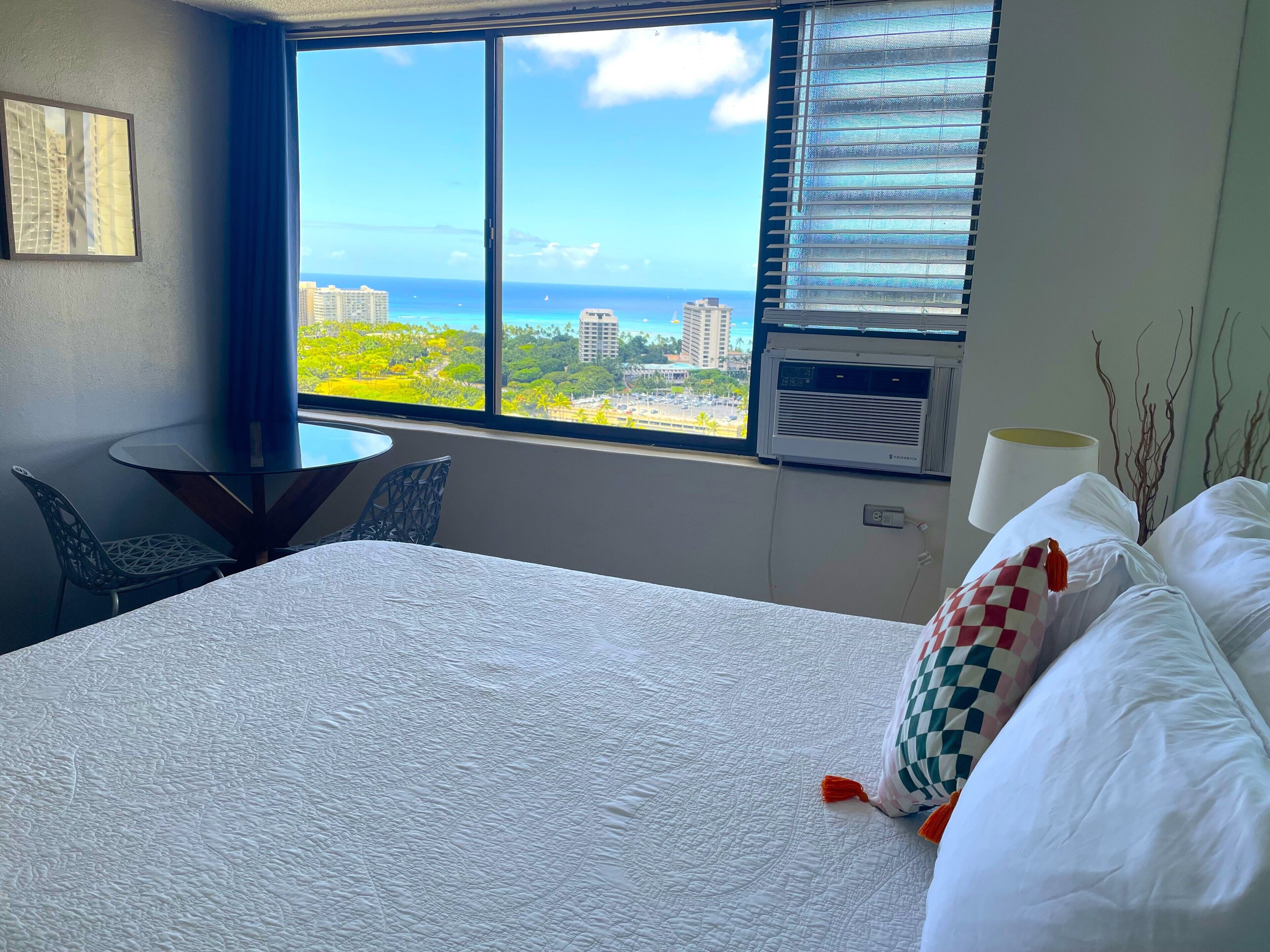 A hotel room with a large bed, a small table, two chairs, a window with a view of the ocean and buildings, and an air conditioning unit.