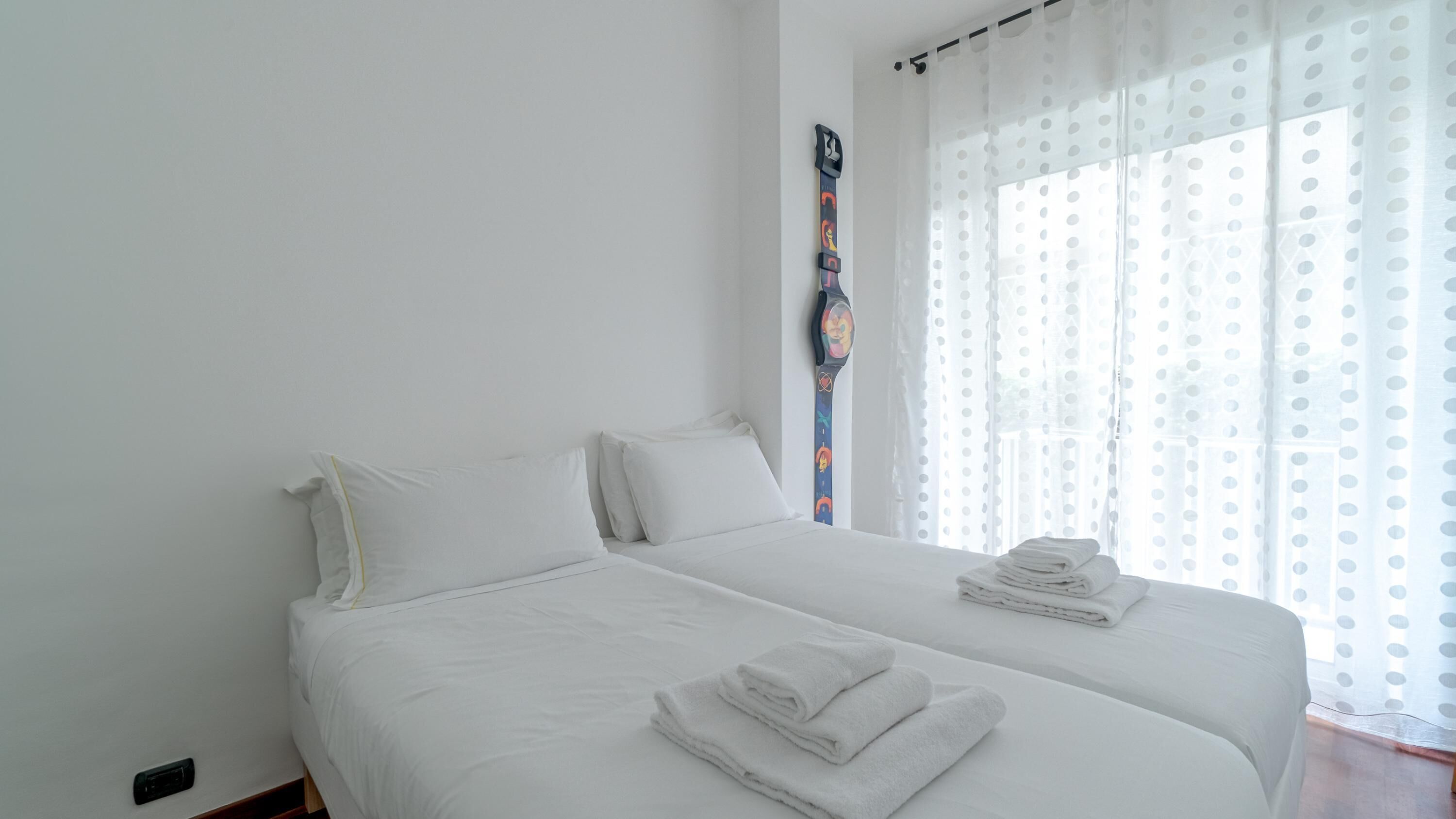 Ein Doppelbett mit weißen Bettwäsche und zwei Kissen, ein Nachttisch mit einem gefalteten Handtuch und ein Fenster mit gepunkteten Vorhängen.