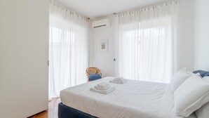 A clean, white bedroom with a large bed, white curtains, a chair, and an air conditioning unit. - Via Aurelia Levante in Rapallo (Rapallo)
