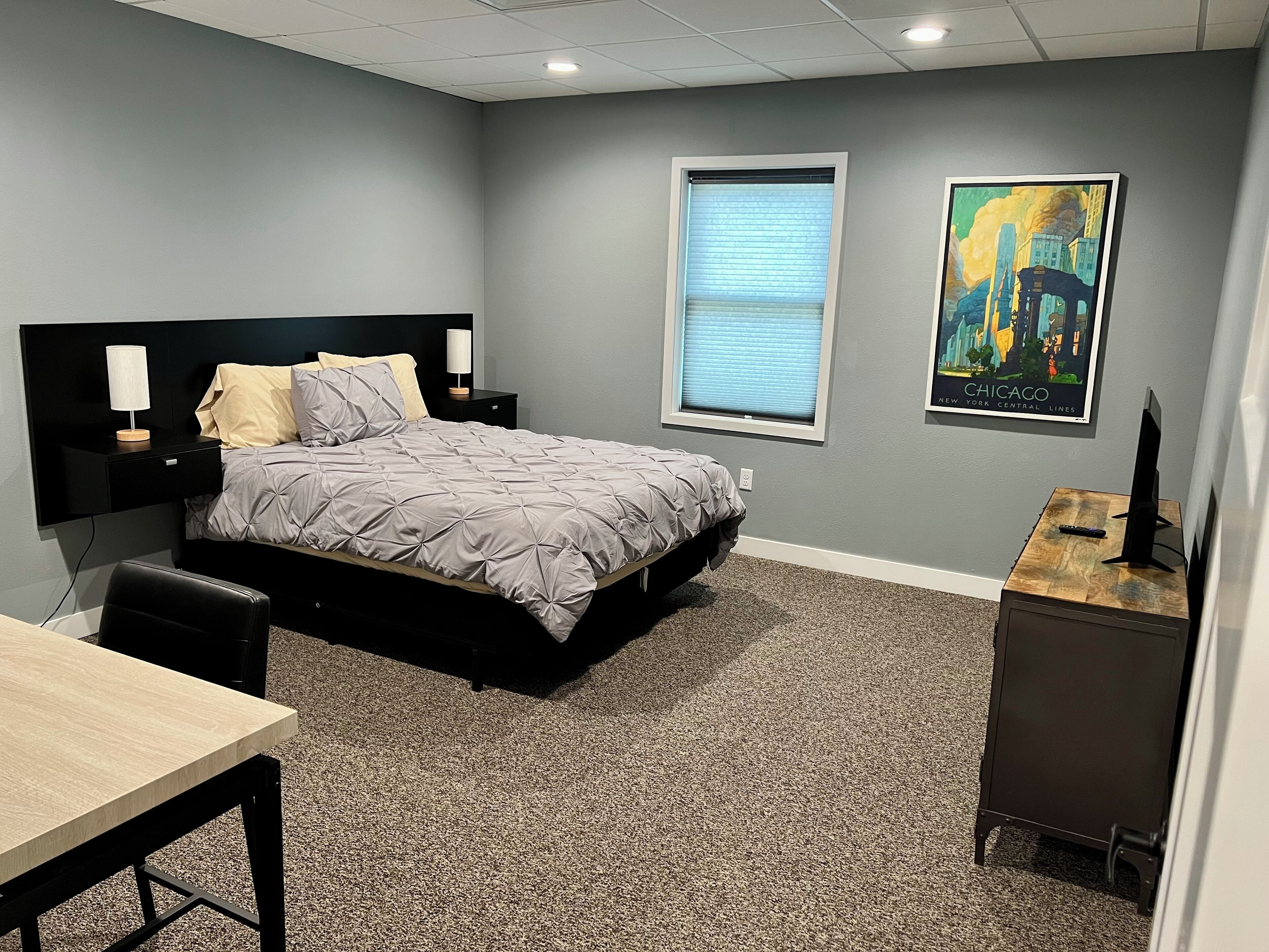 Une chambre à coucher comprenant un lit, des tables de chevet, un bureau, une chaise, une fenêtre avec des stores et un tableau accroché au mur.