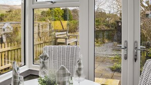A dining area with a table set for four, a wicker chair, and a view of a garden through a glass door. - 2 bedroom accommodation in East Woodburn (East Woodburn)