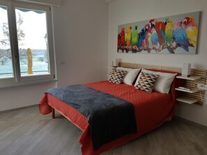 A bedroom with a large bed, a window with a view of the sea, and a colorful parrot painting on the wall. - Mori's Beautiful Sea front Apartment - With A/C (Monterosso al Mare)