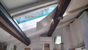 Una habitación con techo inclinado, vigas de madera y un tragaluz. Se observan cuadros enmarcados en las paredes y una lámpara colgante.