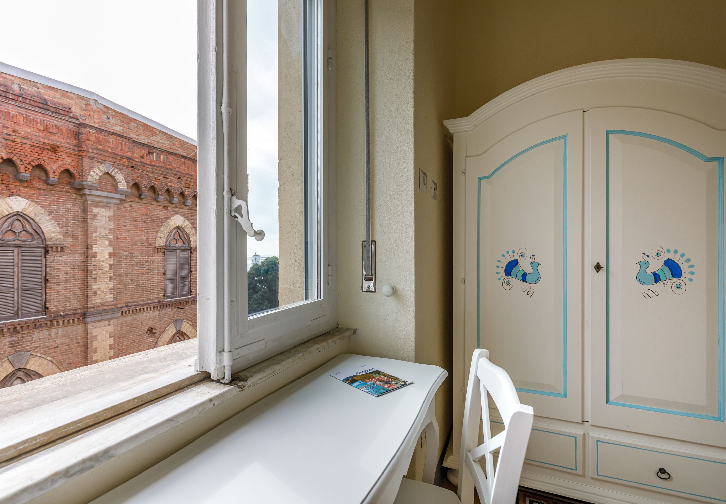 Una habitación con ventana, una mesa blanca, una silla blanca y un armario blanco con diseños decorativos de pavo real.