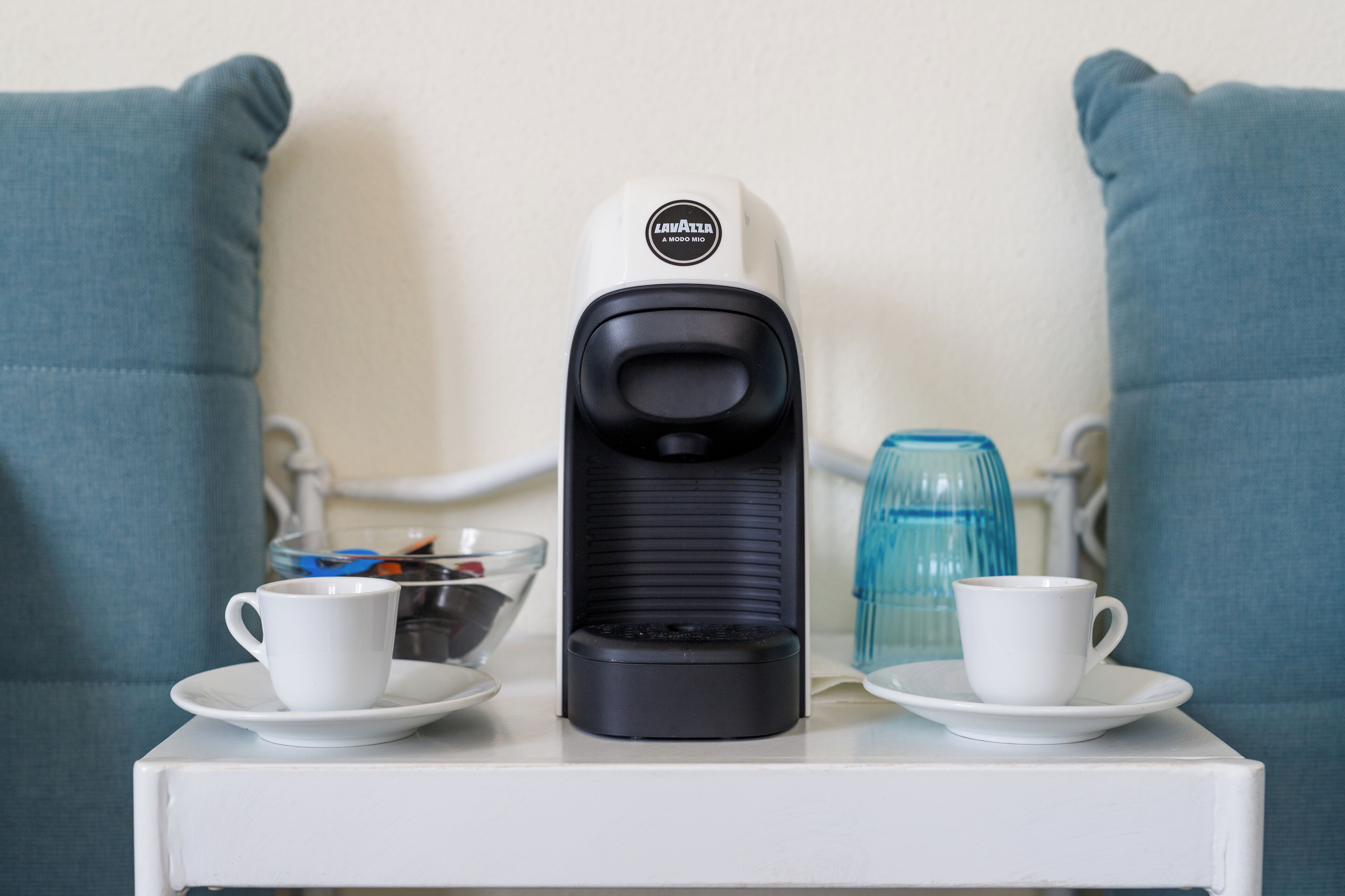 Eine Kaffeemaschine auf einem weißen Tisch, dazu zwei weiße Tassen mit Untertassen, ein Glasbehälter und eine blaue Glasflasche.