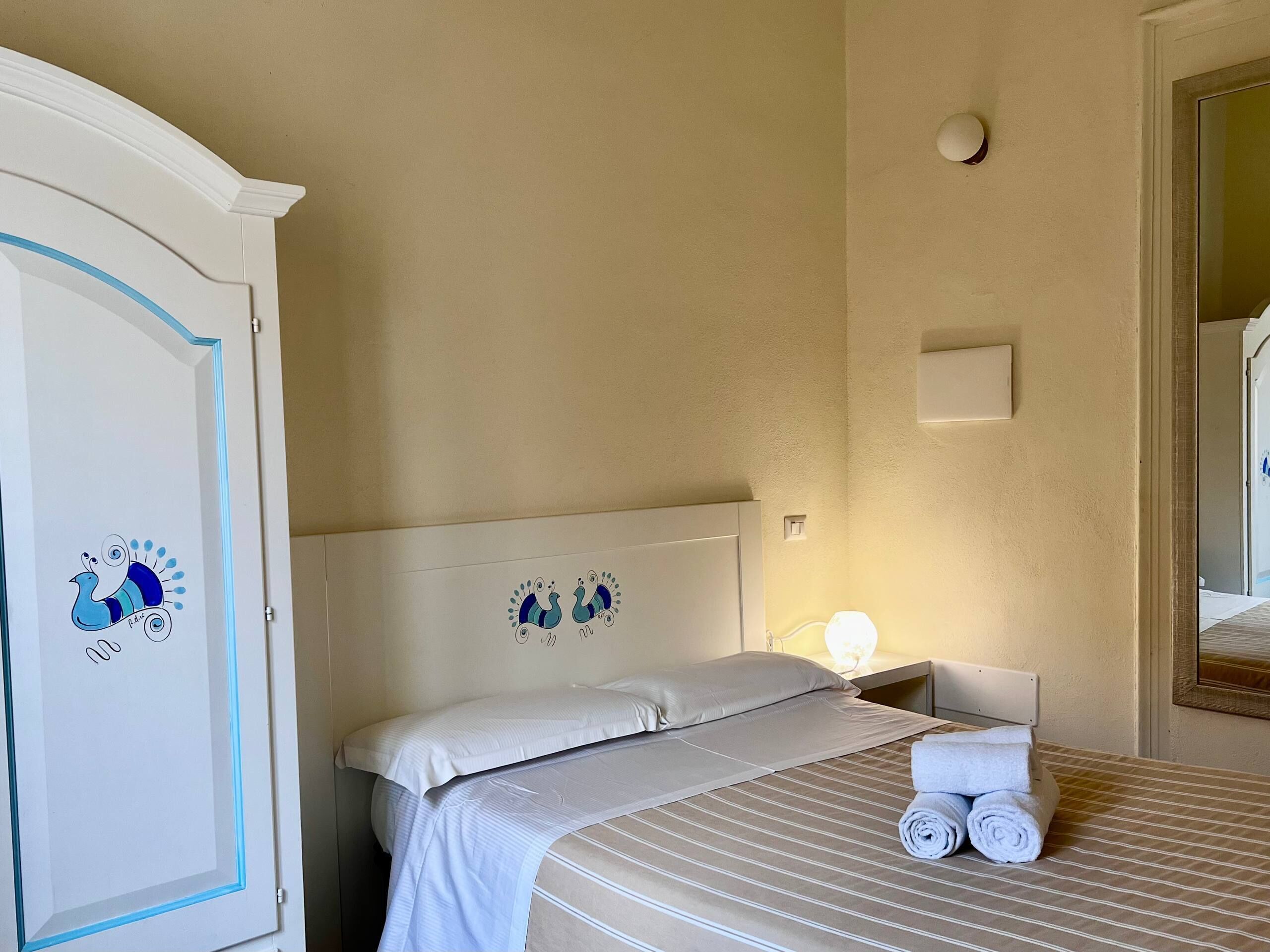 A hotel room with a bed, pillows, a nightstand, a lamp, and a wardrobe.