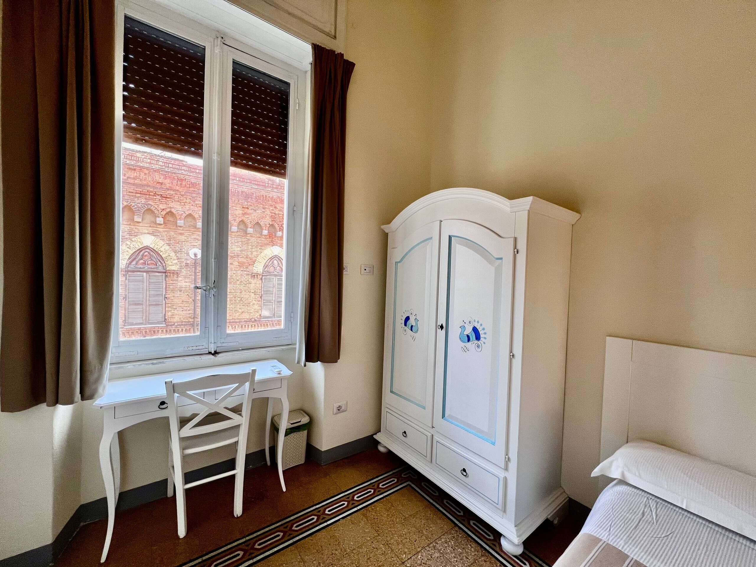 Um quarto com janela, uma escrivaninha branca, um guarda-roupa branco e uma cama.